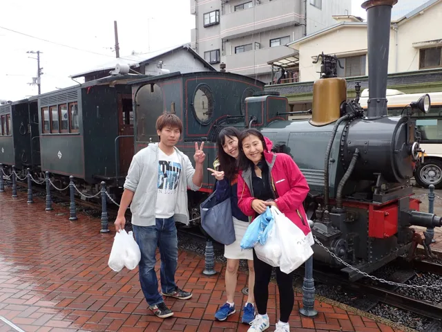 愛媛県松山市・道後温泉周辺（同ツアー写真から推定）での道後温泉周辺と見られる場所で小さな蒸気機関車の前に並ぶ3人の参加者